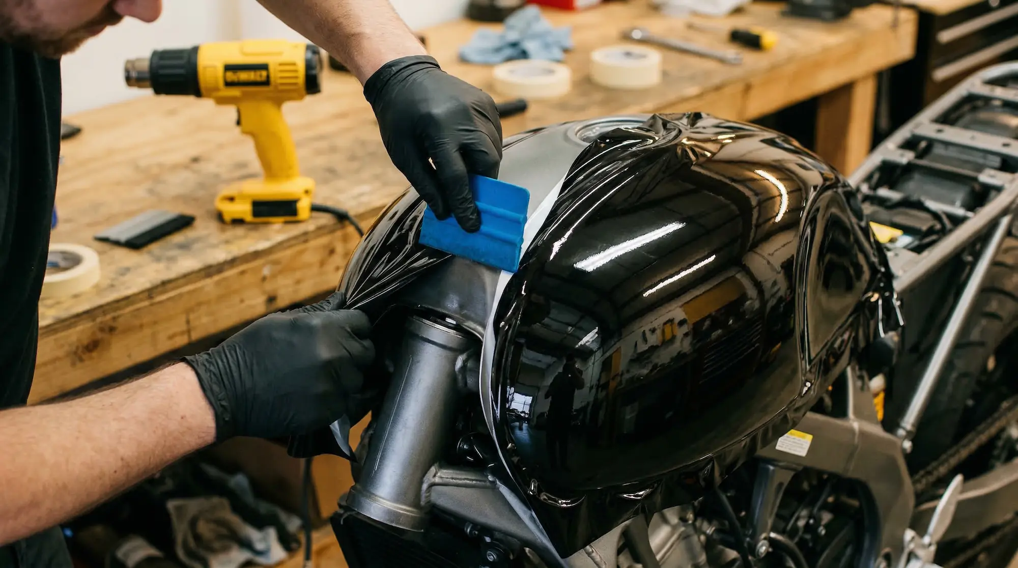 Professional installer applying vinyl wrap to a motorcycle fuel tank using a squeegee and heat gun in a workshop